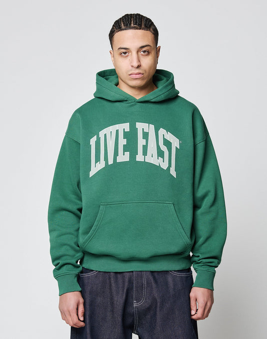 A person faces forward in an oversize fit, green LIVE FAST DIE YOUNG Live Fast College Hooded sweatshirt made from Baumwolle with a bold LIVE FAST frontprint, paired with dark jeans against a plain light background.