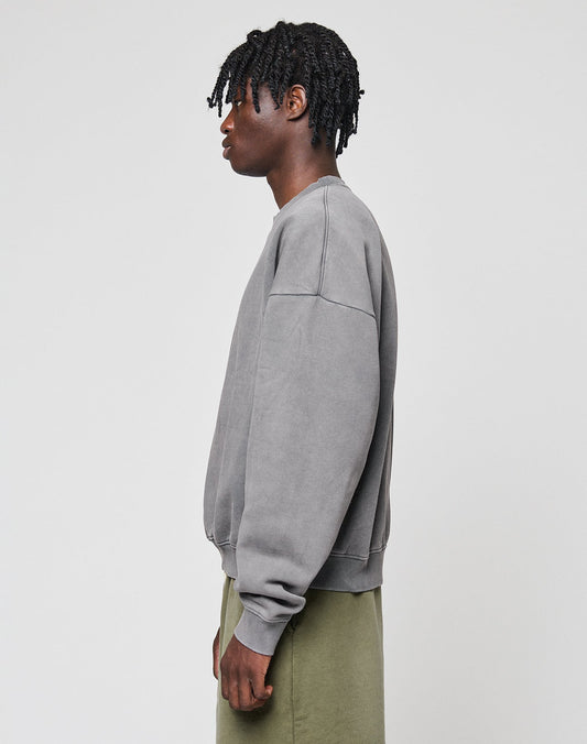 A person with short, twisted hair stands in profile against a plain background, wearing a LIVE FAST DIE YOUNG Basic Blank Sweater in gray and olive green pants.