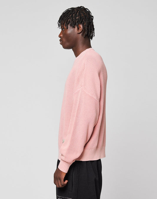 A person with short, twisted hair stands in profile, wearing the LFDY Knit Sweater by LIVE FAST DIE YOUNG and black pants against a plain, light gray background.