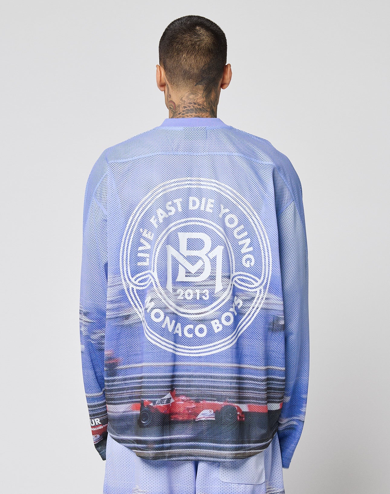 A person with neck tattoos is seen from behind wearing a blue LFDY Yacht Jersey by LIVE FAST DIE YOUNG, featuring an oversize fit, a circular white logo reading LIVE FAST DIE YOUNG MONACO BOYS 2013, and race cars at the bottom.