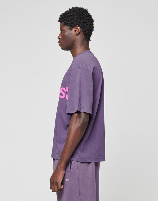 A person stands in profile against a plain background, wearing the LIVE FAST DIE YOUNG Basic OS Livefast Tee in oversize fit purple 100% Baumwolle with a pink frontprint, paired with matching purple shorts.