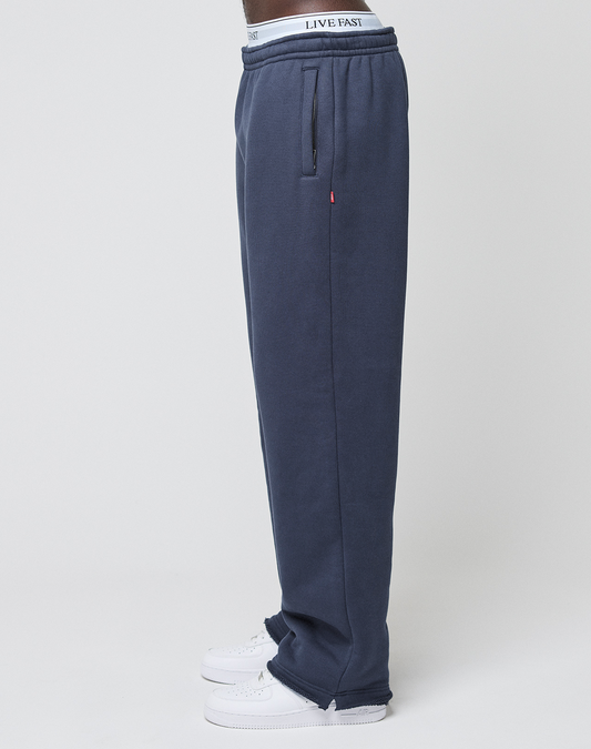 A person stands sideways against a plain white background wearing LIVE FAST DIE YOUNG LFDY Heavyweight Loungepants in navy blue, white sneakers, and underwear with "LIVE FAST" printed on the waistband.