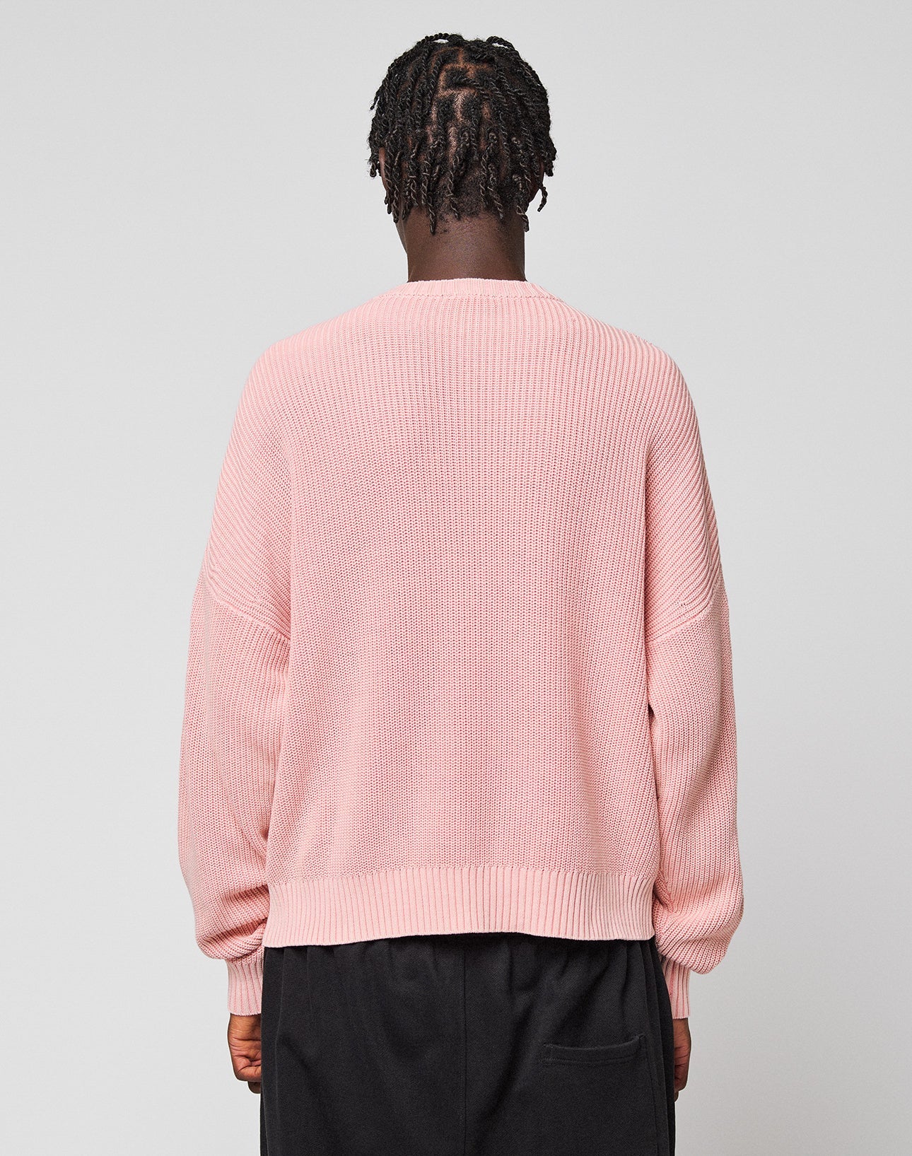 A person with short, twisted hair stands with their back to the camera, wearing a loose-fitting light pink LFDY Knit Sweater by LIVE FAST DIE YOUNG and black pants against a plain light gray background.
