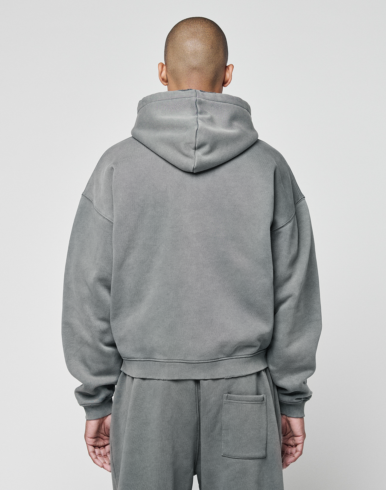 A person with a shaved head faces away from the camera, wearing the LIVE FAST DIE YOUNG Basic Hooded Zip in gray, paired with matching sweatpants against a plain light background.