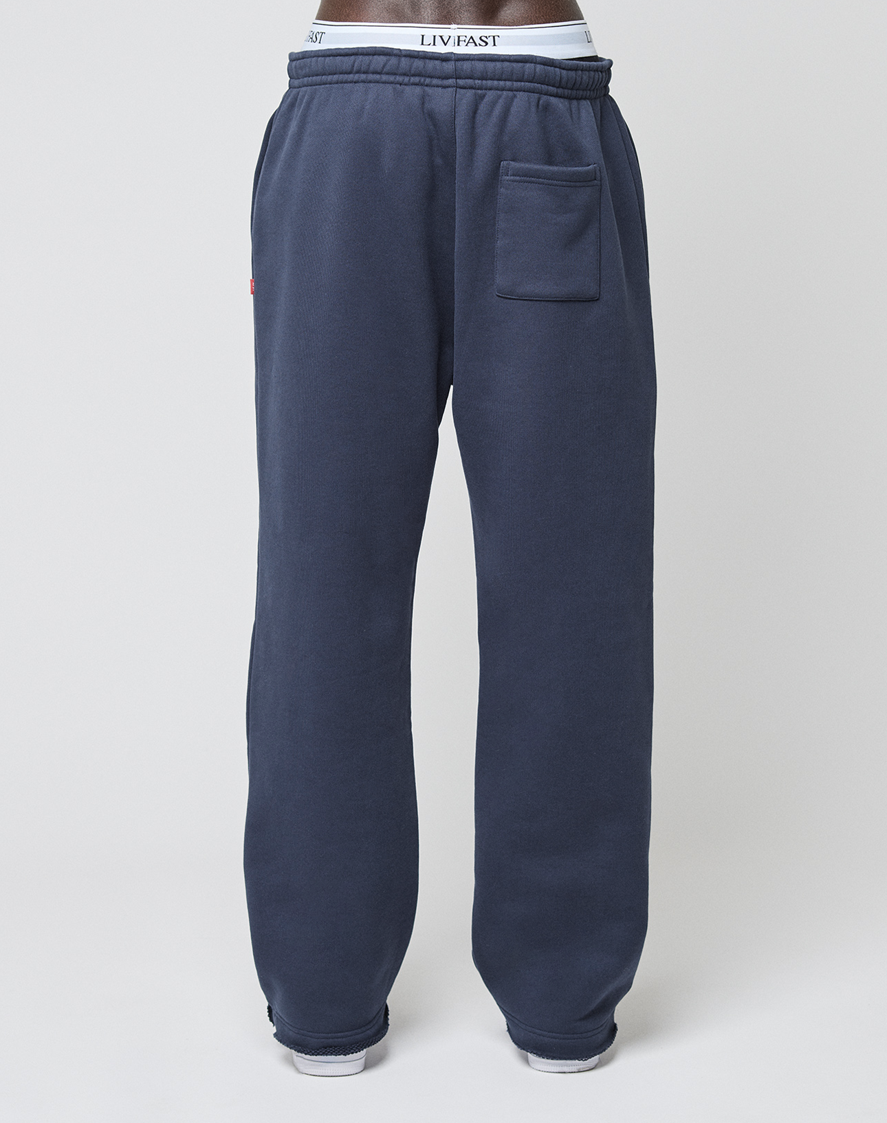 A person wears navy blue LFDY Heavyweight Loungepants by LIVE FAST DIE YOUNG, featuring a patch pocket on the right thigh, white sneakers, and a visible white LIV FAST waistband against a plain white background.