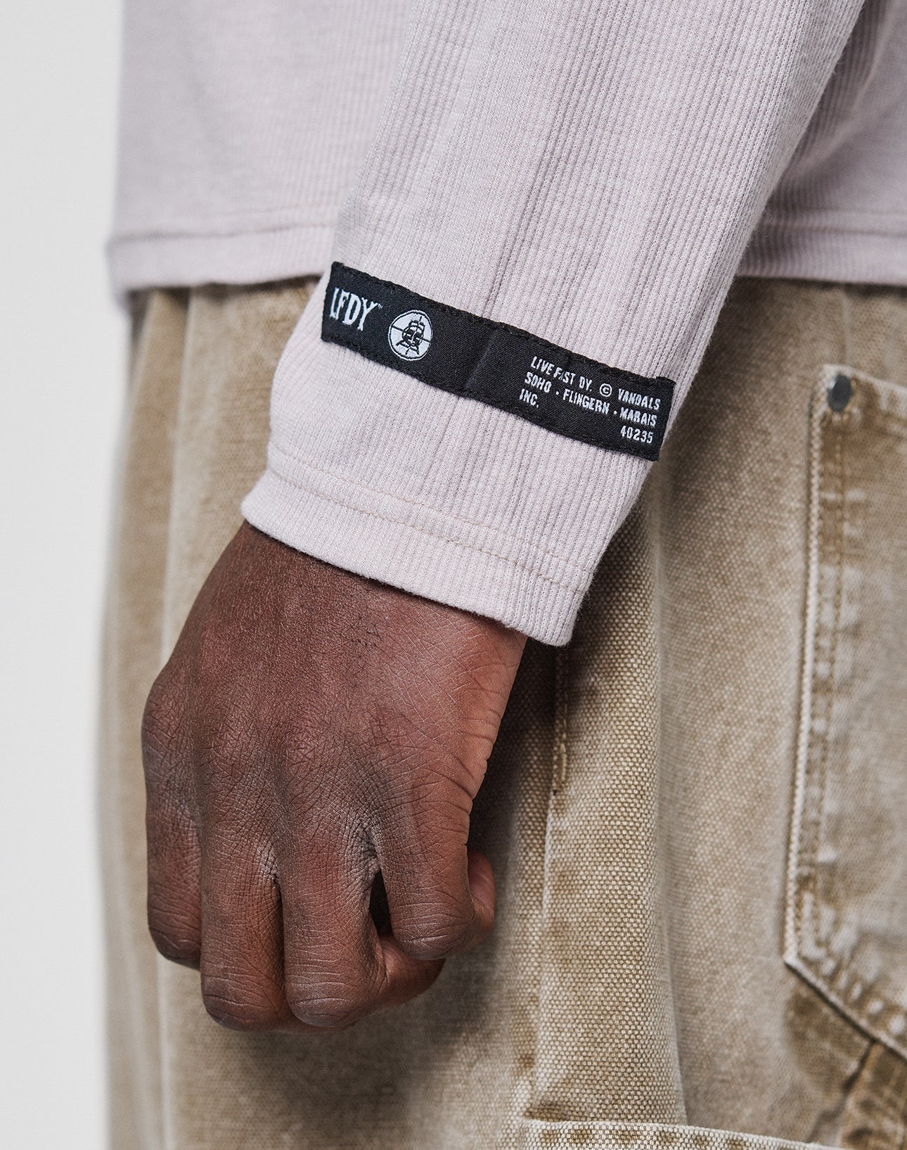 A close-up of a left arm and hand wearing the LIVE FAST DIE YOUNG LFDY Rib Longsleeve in light beige, with a black and white cuff label, detailed topstitching, and paired with light brown pants showing a back pocket seam.
