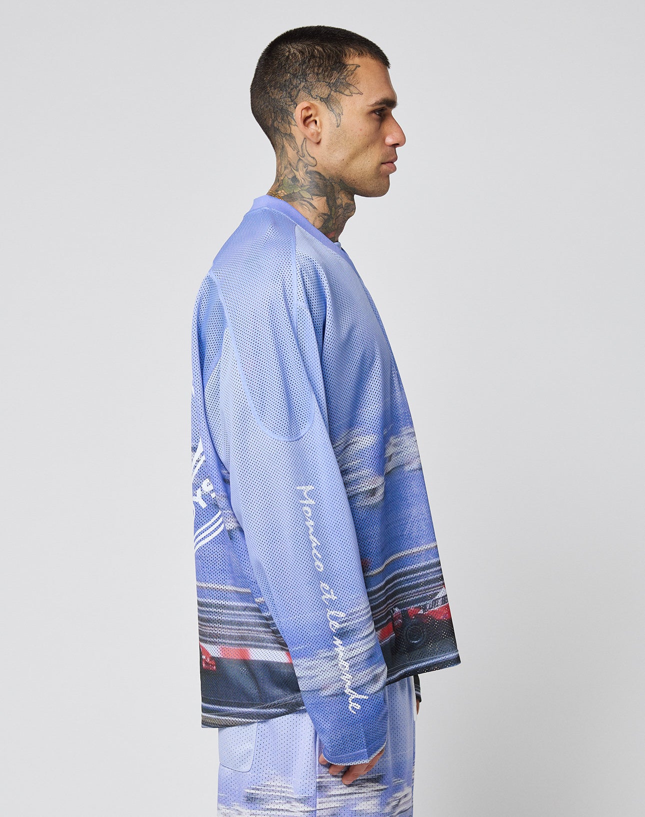 A man with a buzz cut and neck tattoos stands in profile wearing the LIVE FAST DIE YOUNG LFDY Yacht Jersey—an oversize blue and purple graphic long-sleeve shirt with matching shorts, white script on the sleeve—set against a plain light gray background.