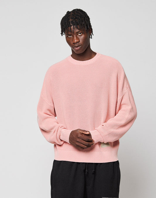 Wearing the LIVE FAST DIE YOUNG LFDY Knit Sweater in light pink with an oversized, ribbed design and paired with black pants, a person stands against a plain light gray background with hands clasped in front.