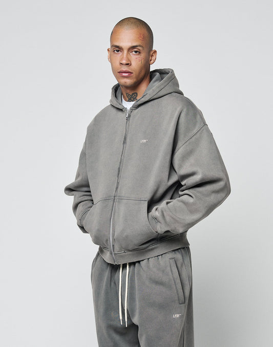 A person with a shaved head and face tattoos models the LIVE FAST DIE YOUNG Basic Hooded Zip in gray—a lightly cropped sweatshirt with subtle logo—and matching sweatpants, hands in pockets, against a plain light background.