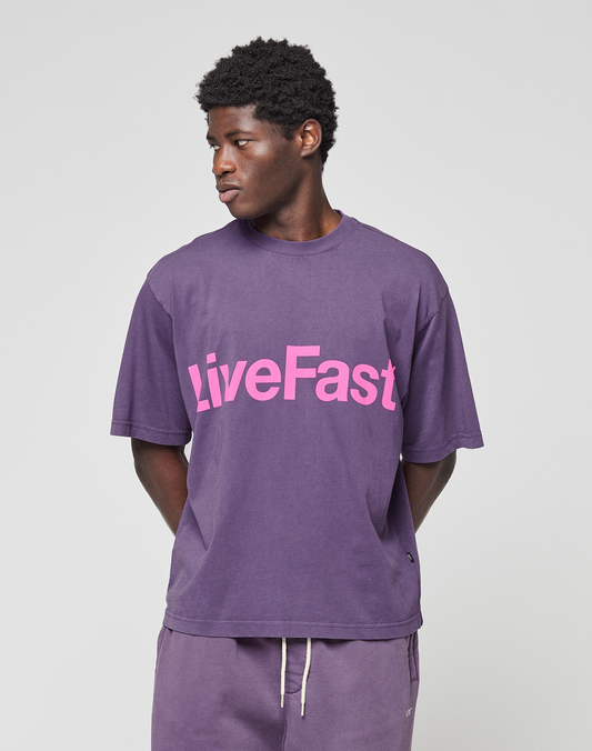 A person stands against a plain background, wearing the LIVE FAST DIE YOUNG Basic OS Livefast Tee—an oversize purple T-shirt in 100% Baumwolle featuring bold pink 'LiveFast' text—paired with matching purple shorts with white drawstrings, looking to the side.