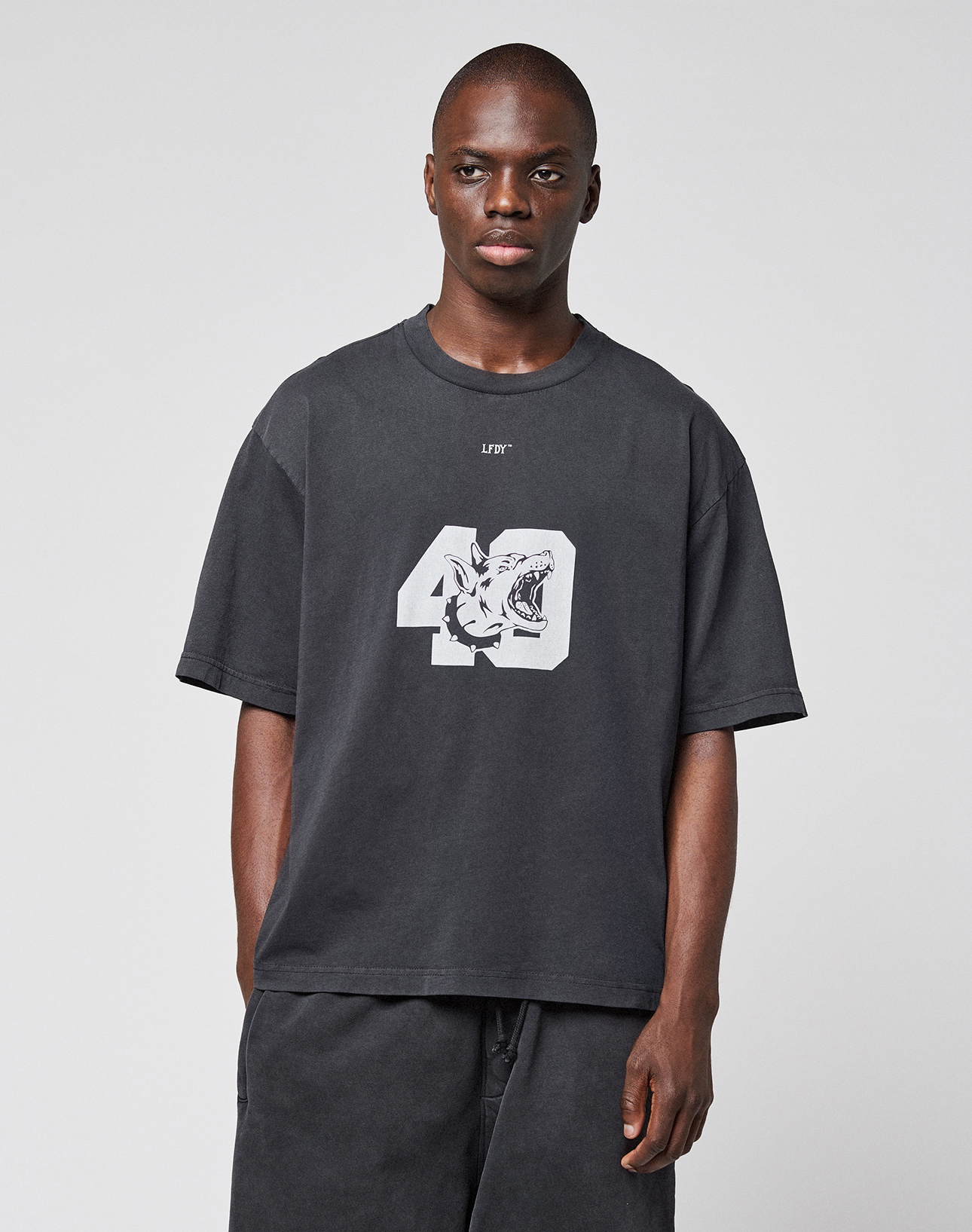 A man with short hair and a neutral expression stands against a light gray background, wearing the LIVE FAST DIE YOUNG LFDY 40 Tee—an oversize black t-shirt made from 100% cotton with a bold 49 and tiger graphic on the front.