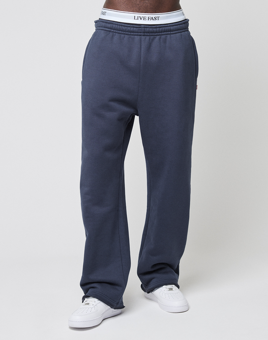 A person wears LIVE FAST DIE YOUNG LFDY Heavyweight Loungepants in dark blue (650gsm, wide fit), white sneakers, and a white waistband with "LIVE FAST" in black, standing against a plain white background. Cropped shoulders down.