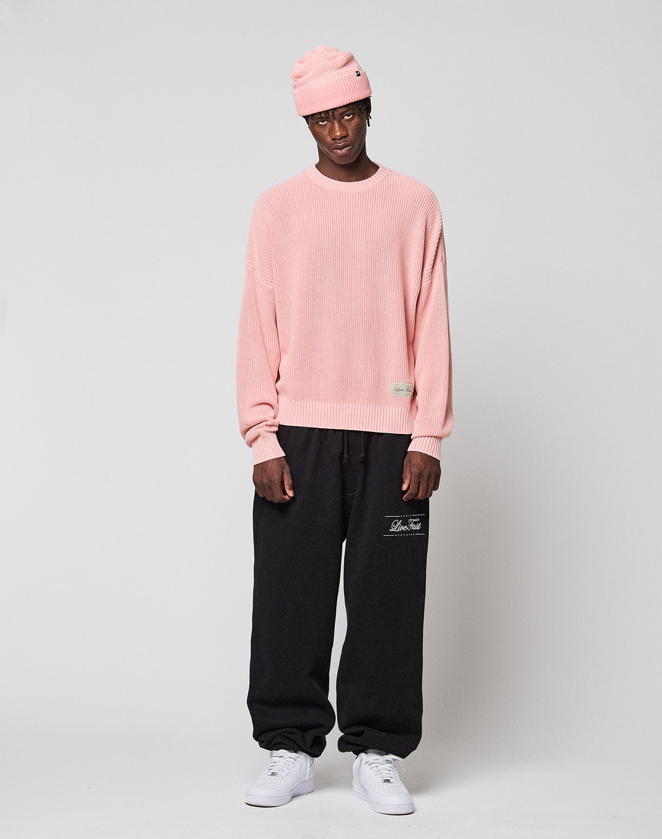 A man stands against a plain background wearing the LFDY Knit Sweater by LIVE FAST DIE YOUNG, a light pink oversize sweater with dropped shoulders, paired with loose black sweatpants featuring white text and white sneakers. He looks at the camera neutrally.
