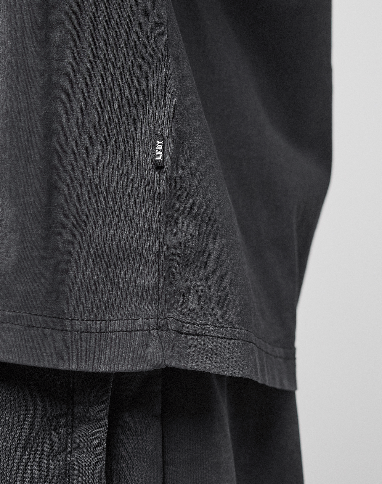 A close-up of the LFDY 40 Tee by LIVE FAST DIE YOUNG, featuring a small black AFRV tag on the seam. Crafted from soft, slightly textured 100% Baumwolle in an Oversize Fit, worn over matching black pants.