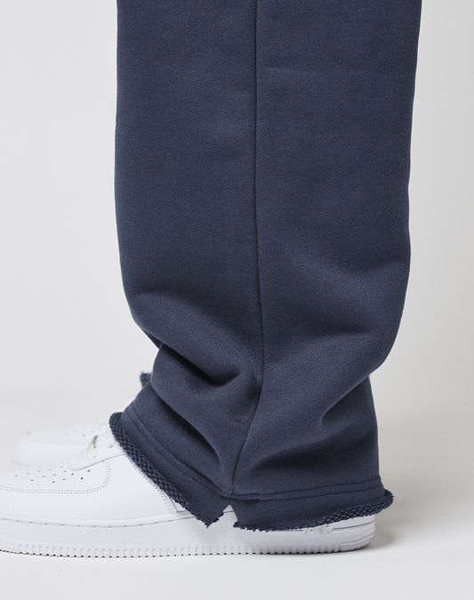 A close-up side view of a person wearing LIVE FAST DIE YOUNG LFDY Heavyweight Loungepants with frayed hems over white sneakers, standing against a plain white background.