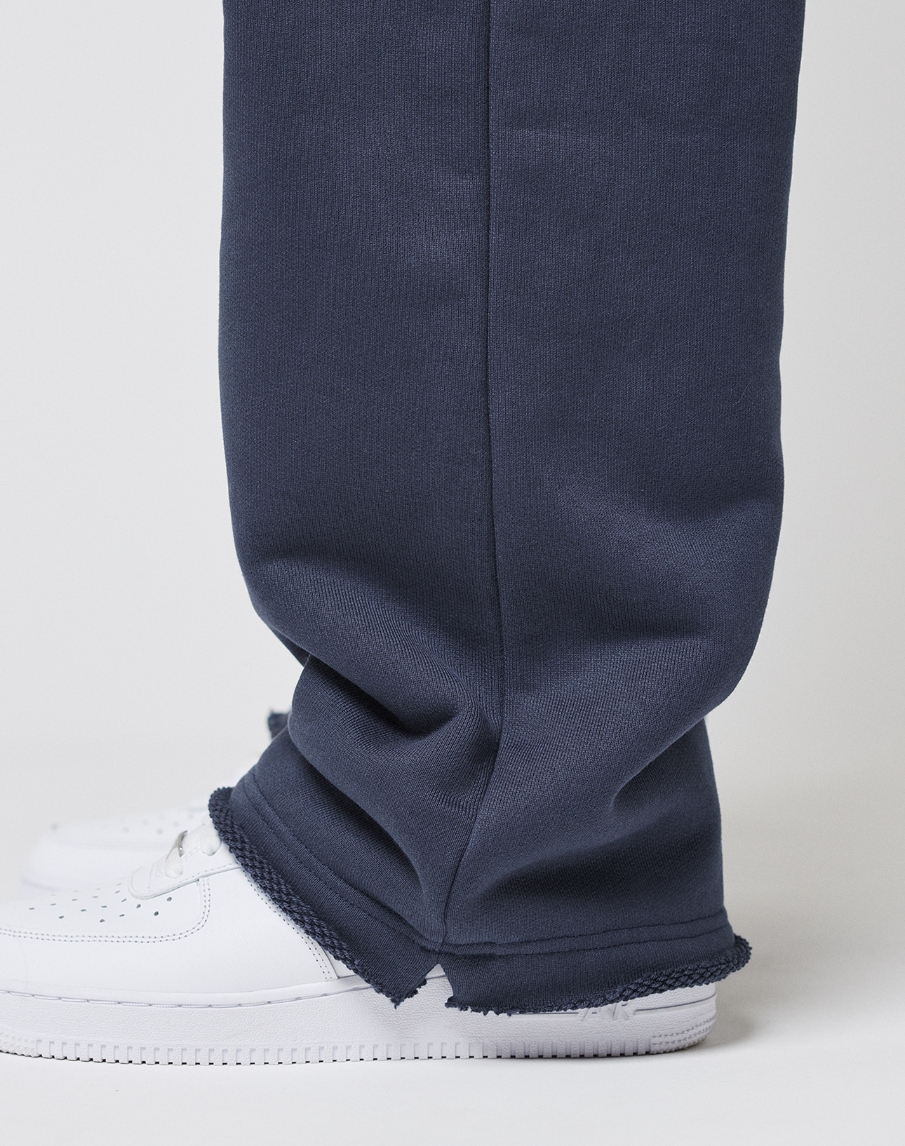 A close-up side view of a person wearing LIVE FAST DIE YOUNG LFDY Heavyweight Loungepants with frayed hems over white sneakers, standing against a plain white background.