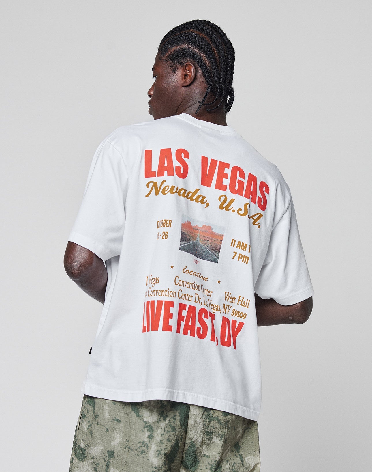 Wearing the Live Fast Vegas Tee by LIVE FAST DIE YOUNG, an oversize white 100% Baumwolle t-shirt with bold red, yellow, and orange LAS VEGAS Nevada, U.S.A. LIVE FAST, DIE YOUNG prints, styled with green camo shorts and braided hair.