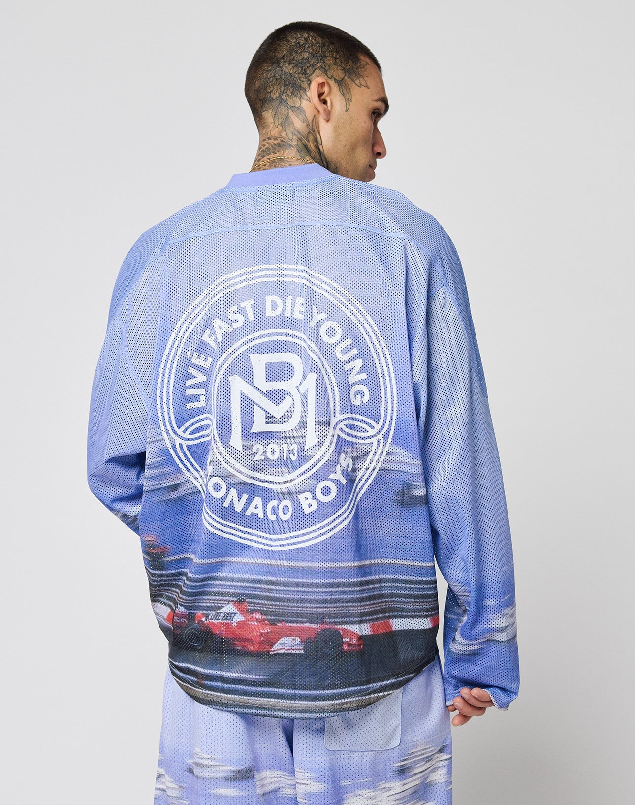 A tattooed, short-haired model wears the LIVE FAST DIE YOUNG LFDY Yacht Jersey—an oversized blue and white long-sleeve shirt featuring racing graphics and a circular LIVE FAST DIE YOUNG MONACO BOYS 2013 print above a race car.