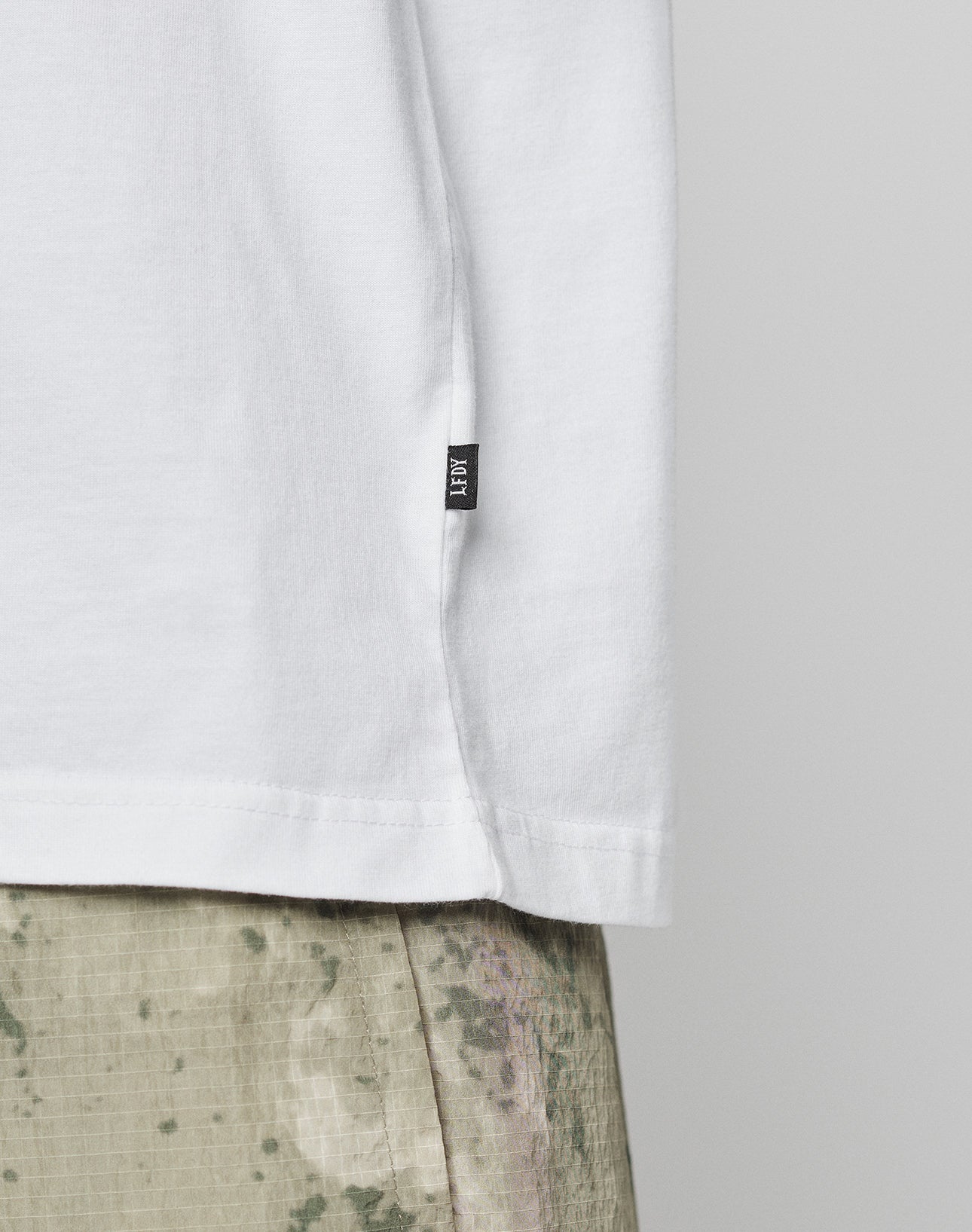 Close-up of the lower part of the LIVE FAST DIE YOUNG Live Fast Vegas Tee in white, made of 100% Baumwolle, featuring a small black side tag, paired with light green and beige camouflage pants.