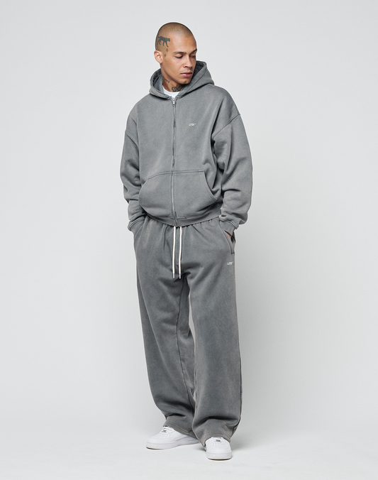 A person stands against a plain background wearing the LIVE FAST DIE YOUNG Basic Hooded Zip—an oversized gray hoodie with matching sweatpants, white drawstrings, and subtle logo prints. Their hands are in their pockets, looking slightly down to the side.