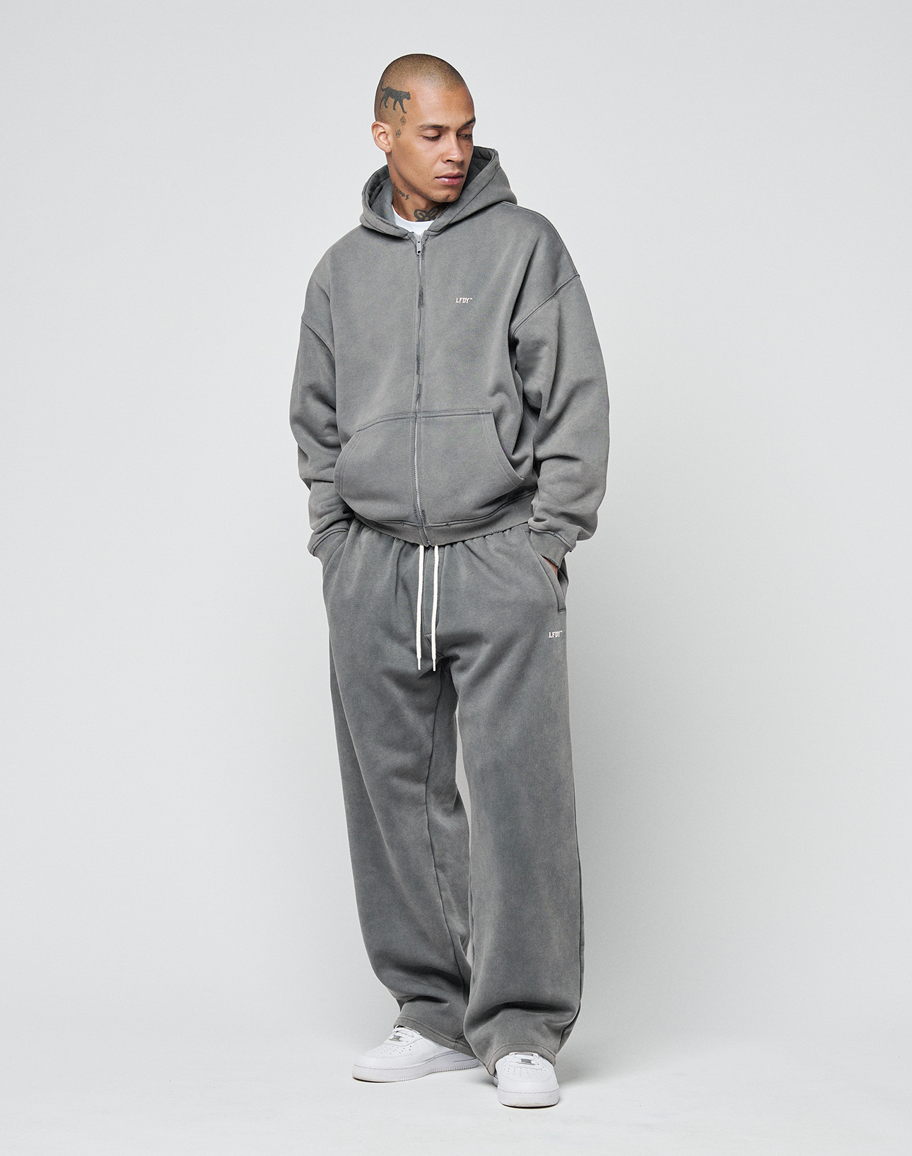 A person stands against a plain background wearing LIVE FAST DIE YOUNG’s Basic Wide Jogger in grey, paired with an oversized zip-up hoodie and white sneakers. Their hands are in their pockets and their expression is neutral.