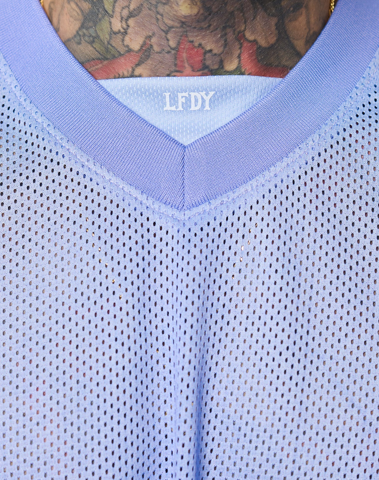 Close-up of a person in the LIVE FAST DIE YOUNG LFDY Yacht Jersey, a lavender mesh shirt with LFDY on the collar, V-neck (V-Ausschnitt), and oversize fit. A floral tattoo is visible on their upper chest and neck. Made from 100% polyester.