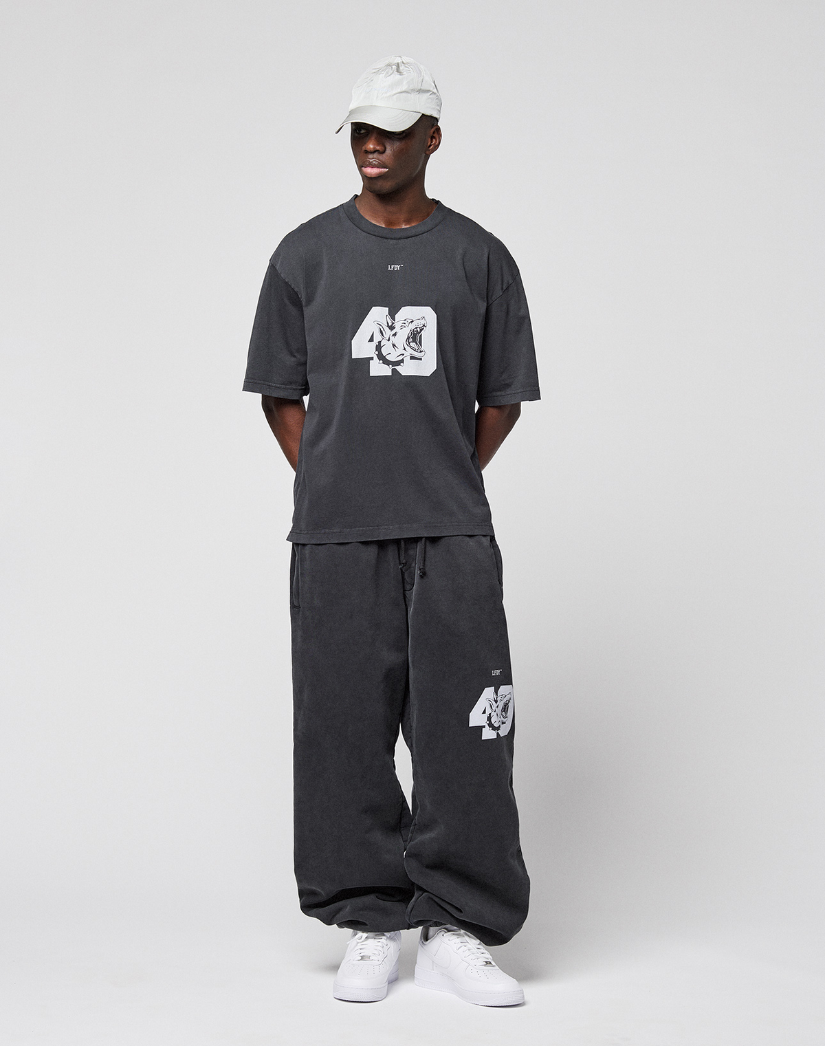 A person in a white cap, oversized dark gray LFDY 40 Tee by LIVE FAST DIE YOUNG with "40" frontprint, matching pants with "40" on the leg, and white sneakers stands against a light background. Hands behind back. 100% Baumwolle.