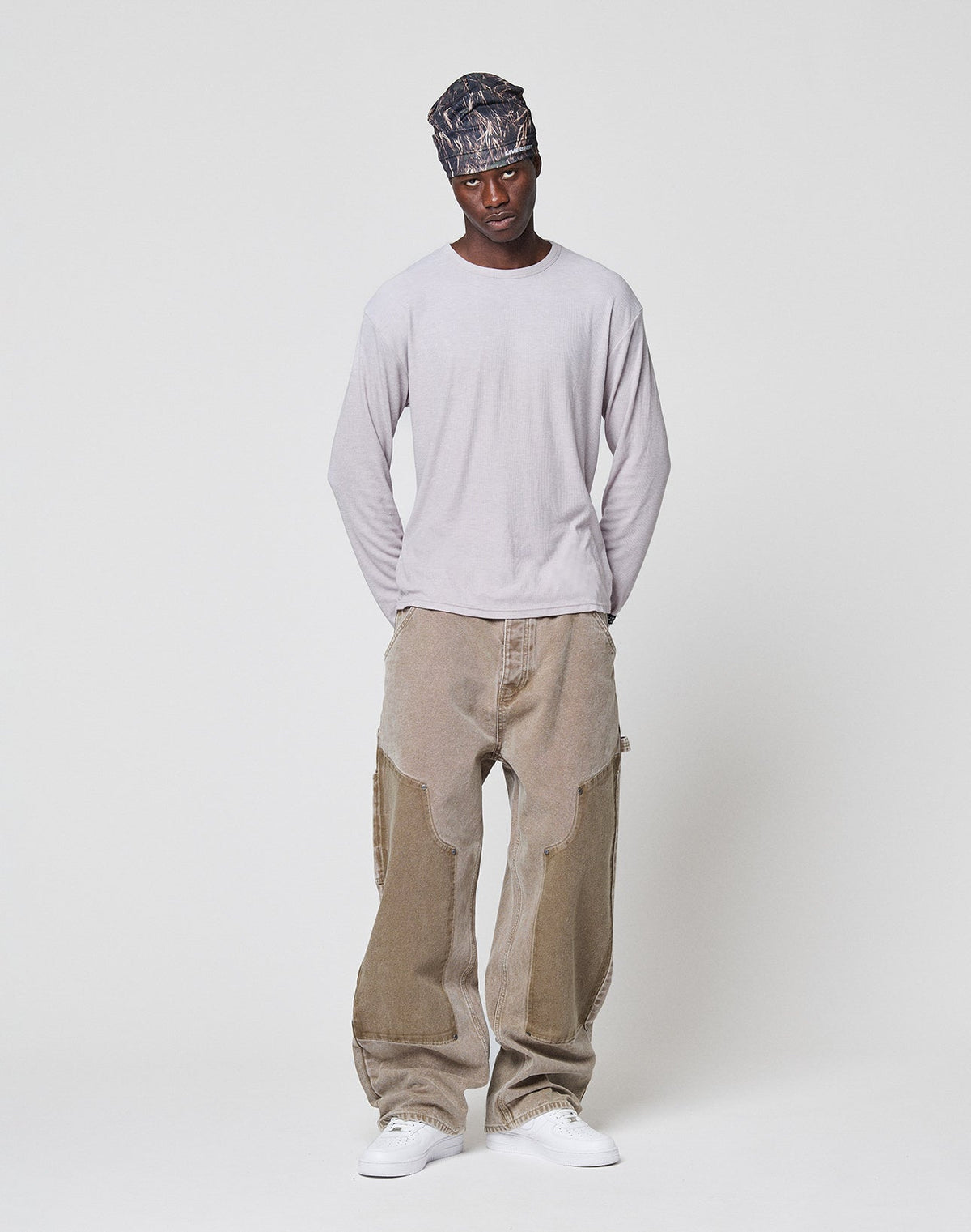 A man in a patterned headscarf, the LFDY Rib Longsleeve by LIVE FAST DIE YOUNG, oversized beige paneled jeans, and white sneakers stands against a plain white background with his hands behind his back.