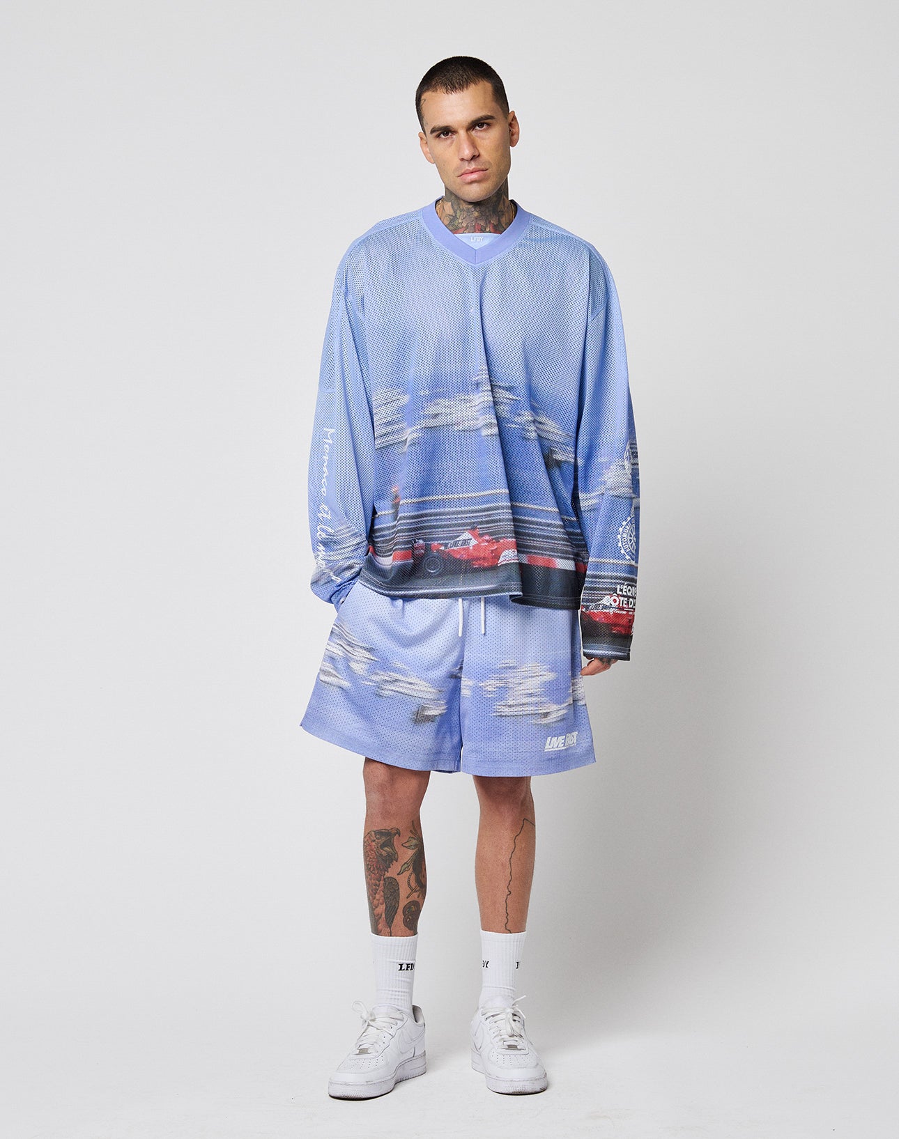 A person with short hair and visible tattoos wears the LFDY Yacht Jersey by LIVE FAST DIE YOUNG, styled oversized with shorts featuring cloud and car graphics, white socks, and white sneakers, standing against a plain white background.