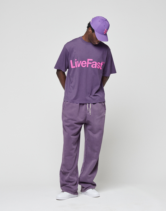 A person wears an oversize fit purple Basic OS Livefast Tee by LIVE FAST DIE YOUNG (100% Baumwolle) with a pink front print, purple cap, and matching sweatpants, standing on a plain white background with hands behind their back.