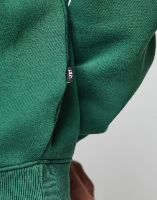Close-up of a person wearing the Live Fast College Hooded by LIVE FAST DIE YOUNG in green, featuring an oversize fit and a small black ACTI label on the side seam. The plain background highlights the fabric and label details.