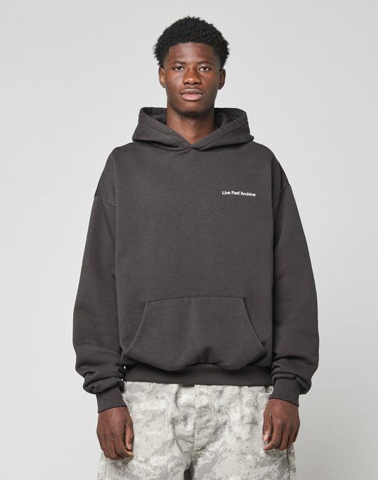 A young man with short, curly hair wears the LFDY Middlefinger Hooded by LIVE FAST DIE YOUNG—a dark, oversize-fit hoodie made from a soft polyester-cotton mix—and light gray camo pants, standing against a plain background with a neutral expression.