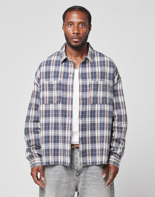 A man wears a LIVE FAST DIE YOUNG Vintage Flannel in blue, white, and gray plaid over a white t-shirt and light gray jeans, standing facing forward against a plain light gray background.