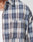 Close-up of a person wearing the LIVE FAST DIE YOUNG Vintage Flannel in blue, white, and brown plaid Oversize Fit over a white ribbed tank. The shirt has a buttoned chest pocket with a small black tag; only neck and chest are visible.