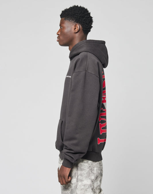 A young man stands in profile wearing the LIVE FAST DIE YOUNG LFDY Middlefinger Hooded, an oversize dark hoodie with red back text, paired with light gray patterned pants made from soft Baumwolle, against a plain light background.