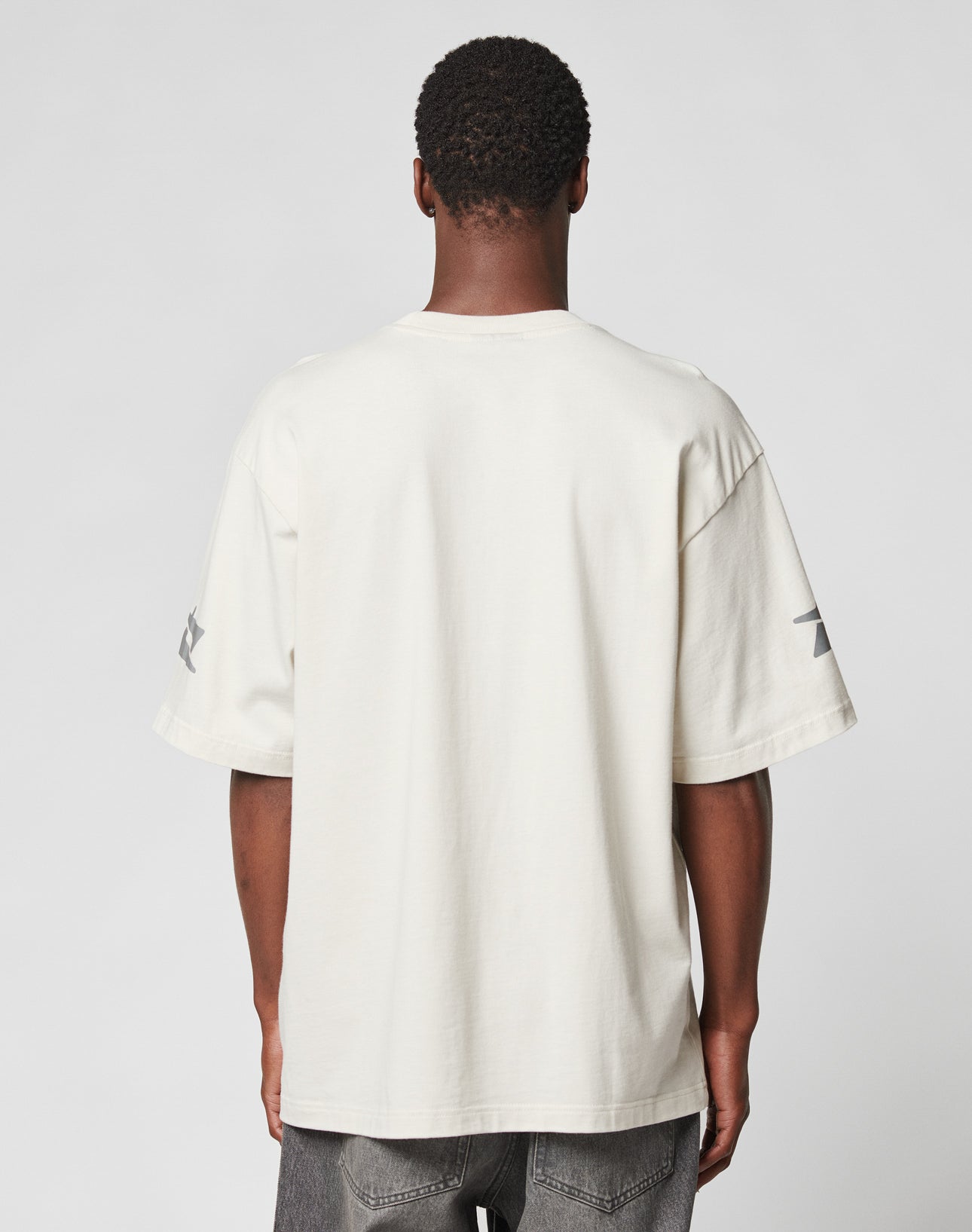 A man faces away from the camera in a LIVE FAST DIE YOUNG LFDY Window Tee—an oversize, off-white 100% Baumwolle shirt with reflective sleeve and logo print—paired with dark gray jeans against a plain gray background.