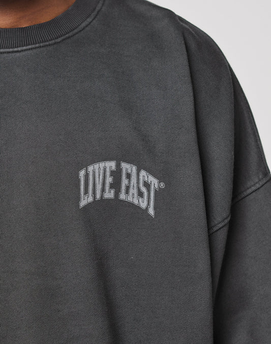 A close-up of a person wearing the Live Fast Berlin Sweater by LIVE FAST DIE YOUNG, featuring a dark gray, oversized fit with LIVE FAST in white uppercase letters on 100% Baumwolle and subtle topstitching details.