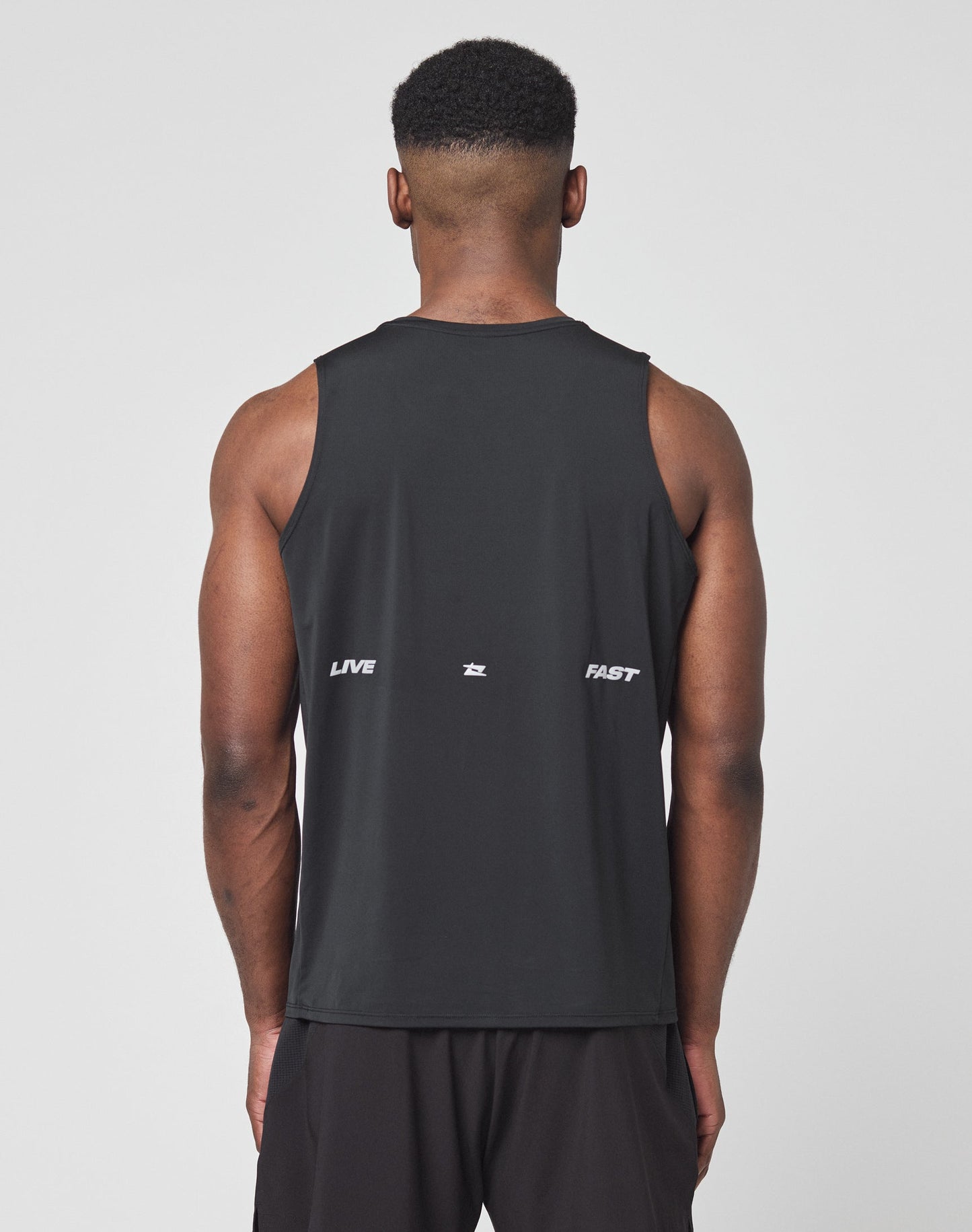 A close-up of a persons back in the Live Fast Performance Tank by LIVE FAST DIE YOUNG, a slim fit black sleeveless nylon-spandex shirt featuring LIVE and FAST in white, divided by a small reflective print at the center.