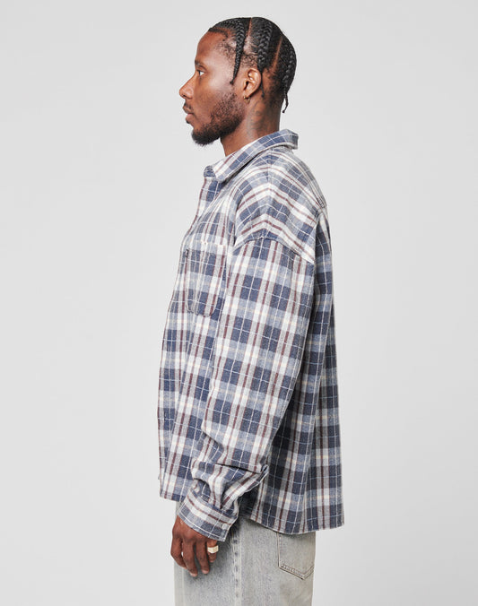 A man in profile against a plain background wears the LIVE FAST DIE YOUNG Vintage Flannel—a blue, white, and gray plaid oversize shirt with Brusttasche—paired with light jeans in Vintage Waschung.