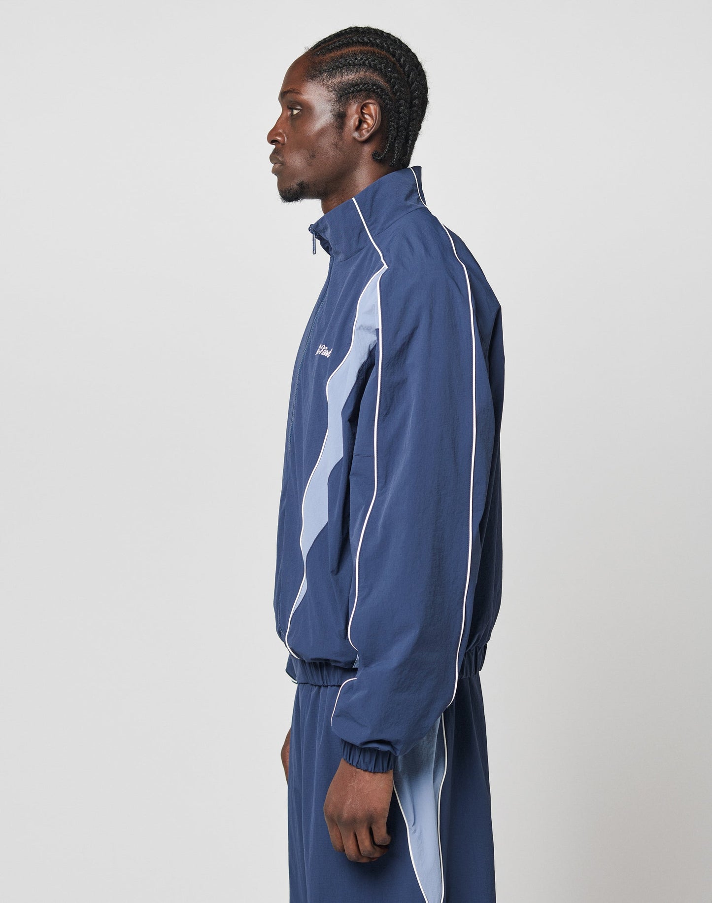 A man with braided hair stands against a plain light background, wearing the LIVE FAST DIE YOUNG Basic Nylon Trackjacket in navy blue with light blue and white stripes and elastische Bündchen, arms relaxed at his sides.