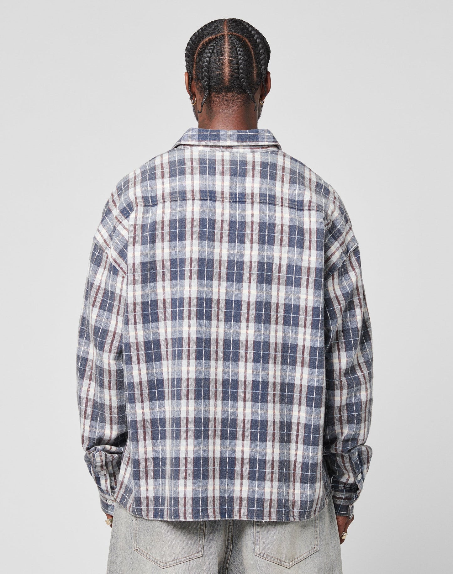 A person with braided hair is seen from behind, wearing an oversize fit LIVE FAST DIE YOUNG Vintage Flannel in blue, white, and gray plaid with a Brusttasche and light-wash jeans against a plain light gray background.