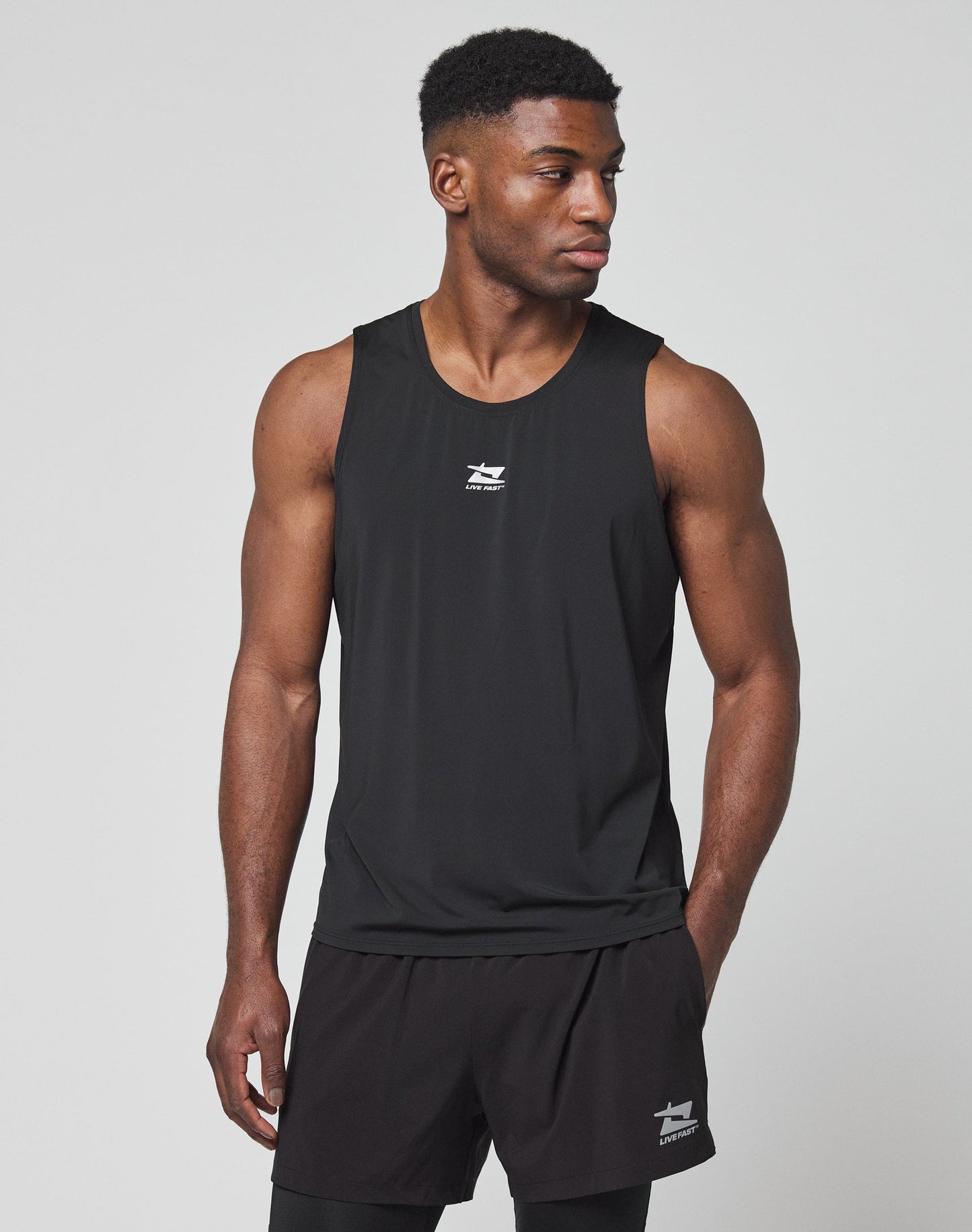 A man stands in profile against a plain white background, wearing the LIVE FAST DIE YOUNG Live Fast Performance Tank and black shorts. His arms are relaxed by his sides, and he looks straight ahead.