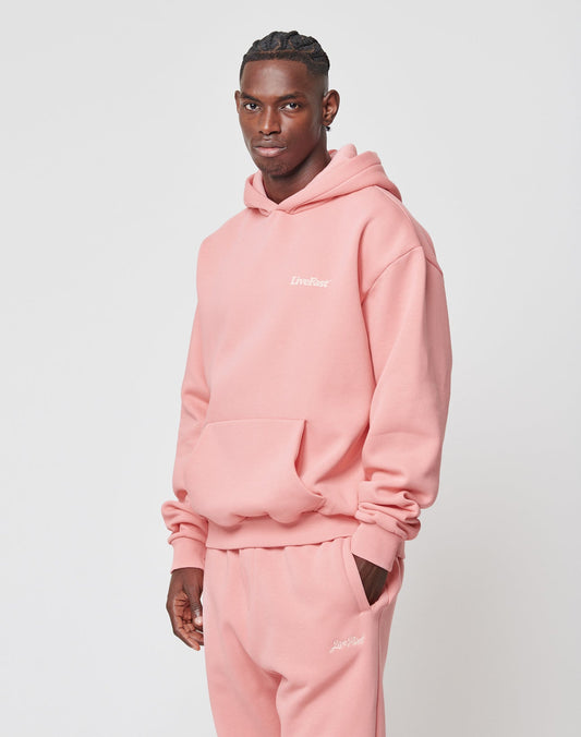 A man in a light pink LIVE FAST DIE YOUNG Basic 360 Hooded oversize fit hoodie and sweatpants stands against a plain background, showing topstitching, a front pocket, and white chest text. He looks at the camera neutrally.