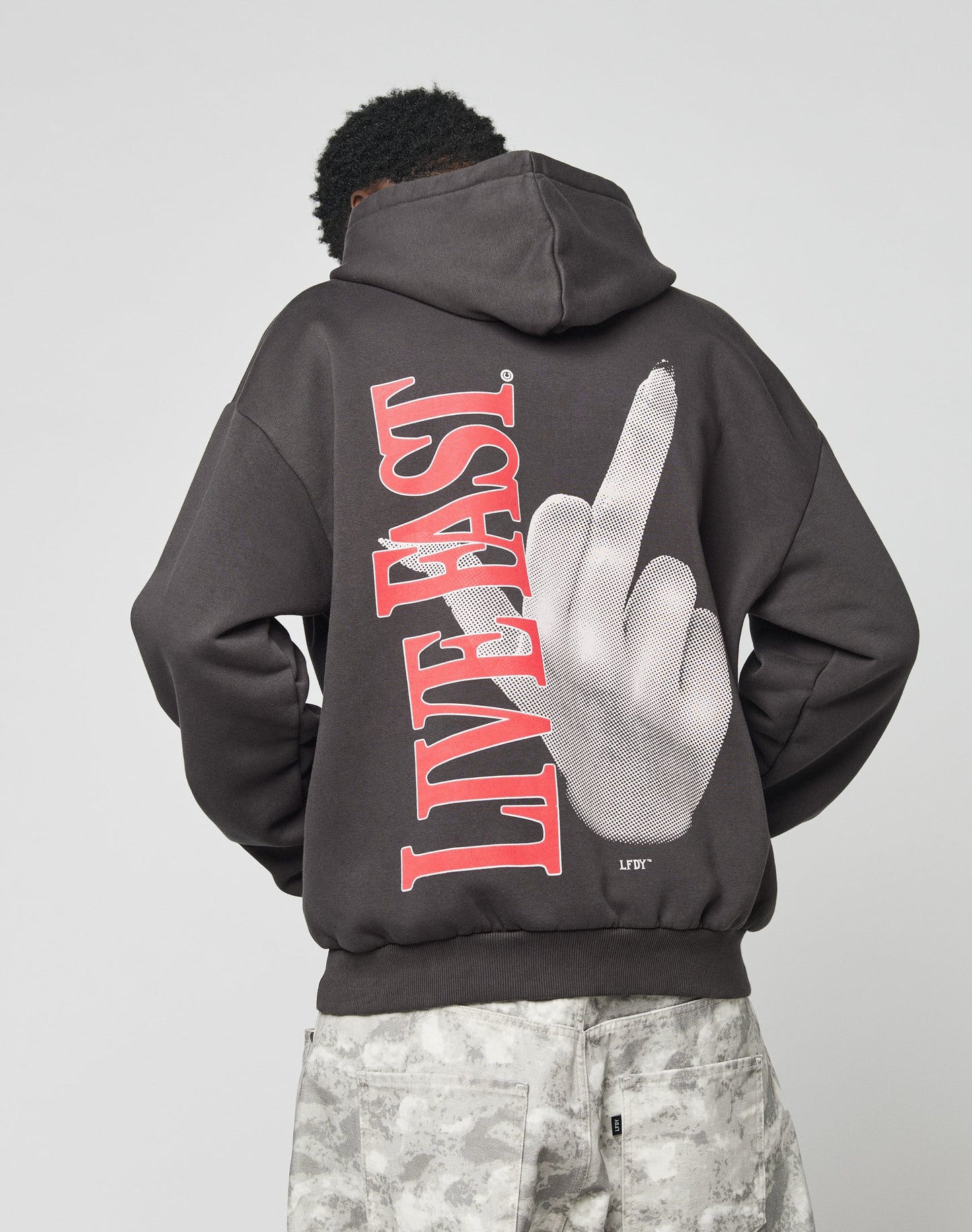 A person in an oversize black LFDY Middlefinger Hooded by LIVE FAST DIE YOUNG, featuring a large hand middle finger graphic and bold red LIVE FAST text on the back, faces away from the camera wearing light gray camo pants.
