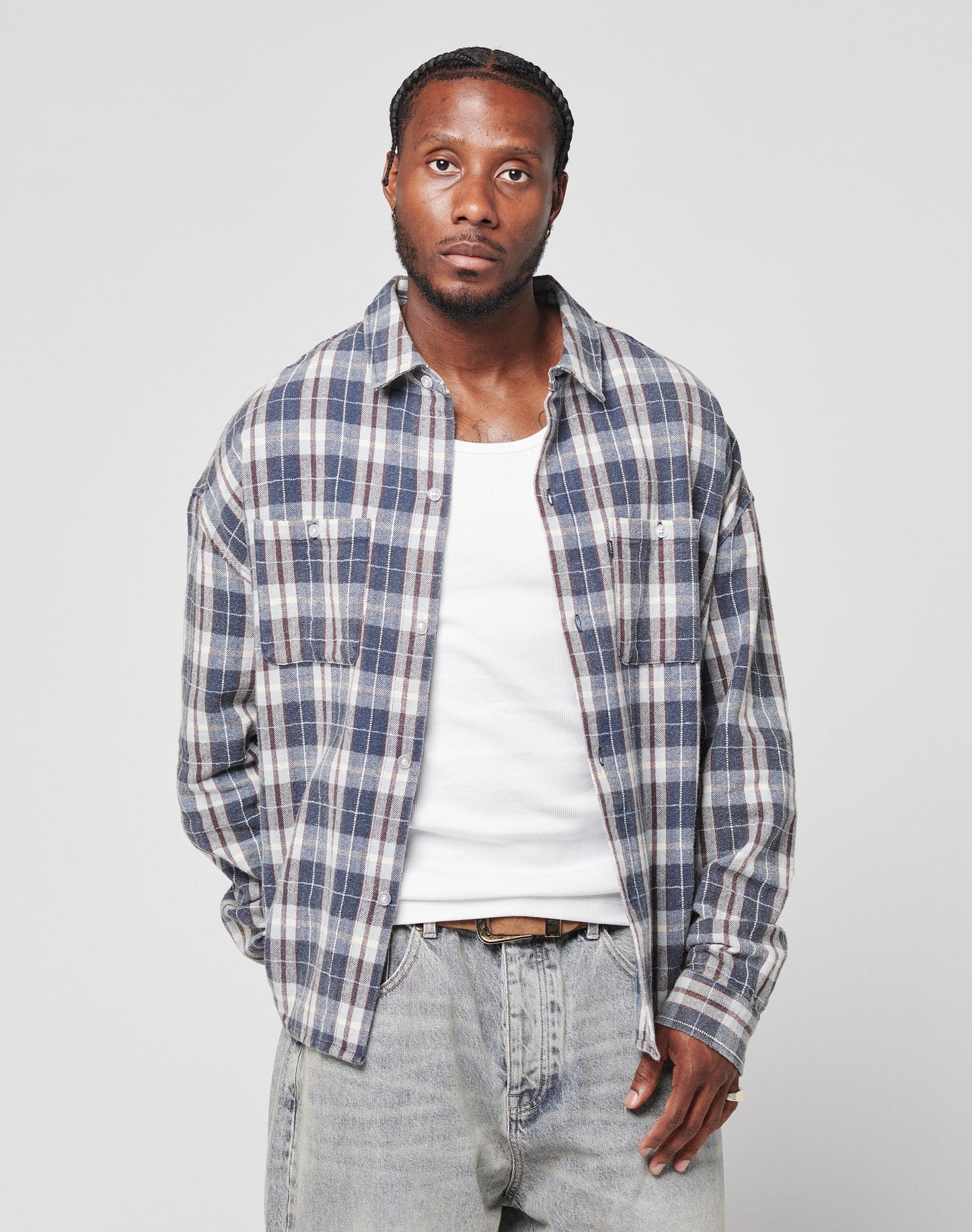 A man with short, braided hair wears the LIVE FAST DIE YOUNG Vintage Flannel—an oversize blue and gray plaid shirt with a chest pocket—over a white tank top and light-wash jeans, standing against a plain light gray background.