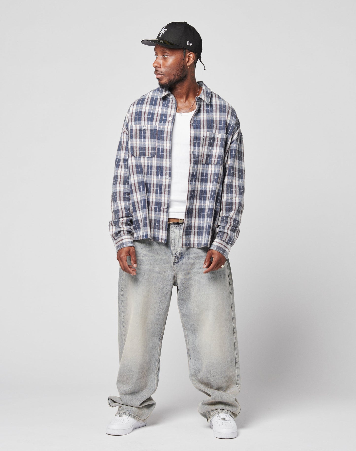 A man wears a LIVE FAST DIE YOUNG Vintage Flannel shirt over a white tee, with baggy light-wash jeans, a black cap, and white sneakers, standing slightly turned against a plain light background.