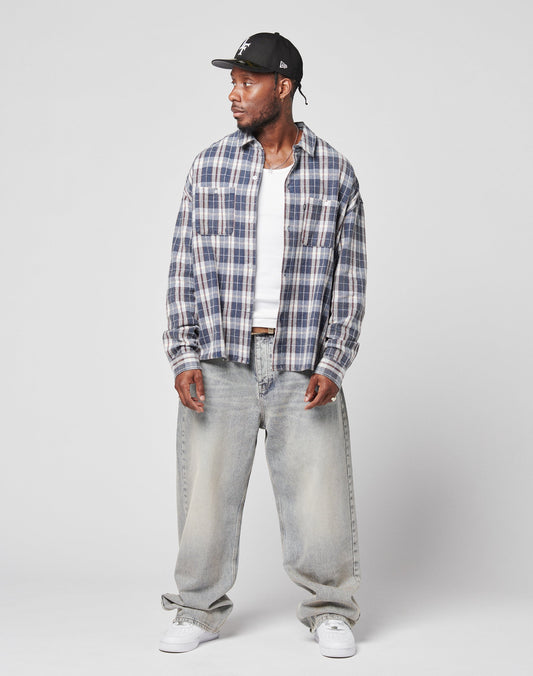 A man in a black cap, blue and white plaid shirt over white tee, LIVE FAST DIE YOUNG LFDY Baggy Denim jeans (baggy fit, faded, wide straight leg), and white sneakers stands slightly turned against a plain gray background.