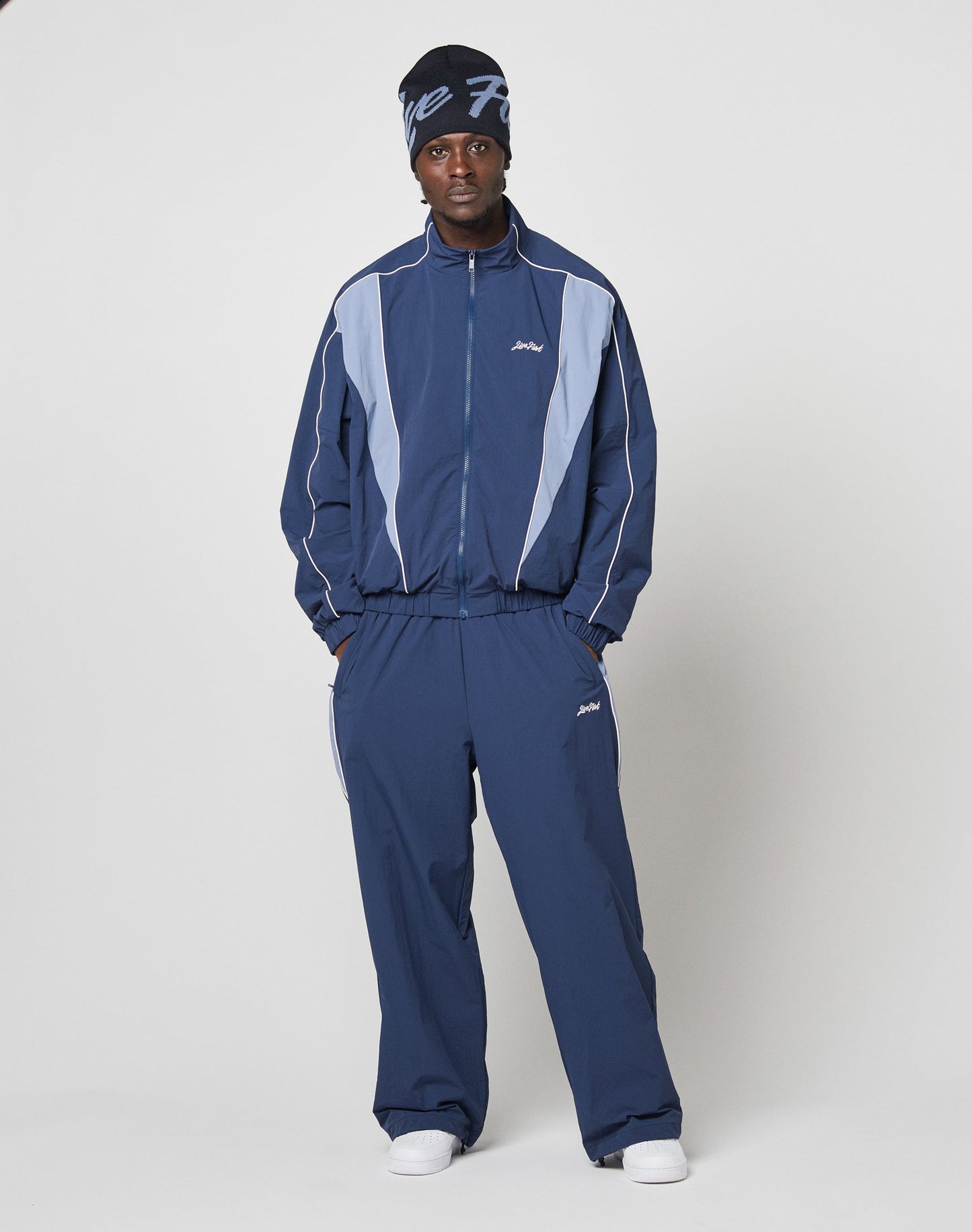 Wearing LIVE FAST DIE YOUNG Basic Nylon Trackpants with Reißverschlusstaschen and Elastikbund, paired with a matching beanie featuring white text and white sneakers, a person stands against a plain backdrop, hands in pockets, looking at the camera.