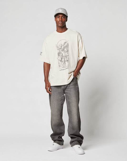 A man stands against a plain background wearing the LFDY Window Tee from LIVE FAST DIE YOUNG, an oversize beige graphic t-shirt with reflective sleeve and logo print, paired with relaxed grey jeans, a white cap, and sneakers. He looks neutral with one hand in his pocket.