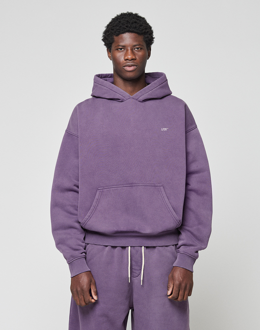 A young man faces forward in a purple LIVE FAST DIE YOUNG Basic Evidence Hooded Oversize Fit hoodie and matching sweatpants. Made from a soft Baumwolle Polyester blend, the hoodie has a small white logo on the left chest. The background is light and plain.